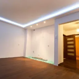 Jasne wnętrze po remoncie z drewnianą podłogą, listwami LED na suficie i widokiem na sąsiednie pomieszczenie przez otwarte drzwi.