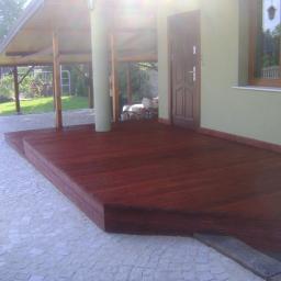 FHU Jarko - Ciemnoczerwony taras z drewna przy domu z zieloną elewacją. Widoczny fragment pojazdu z lewej strony. Zadaszenie tarasu wsparte na drewnianych słupach.