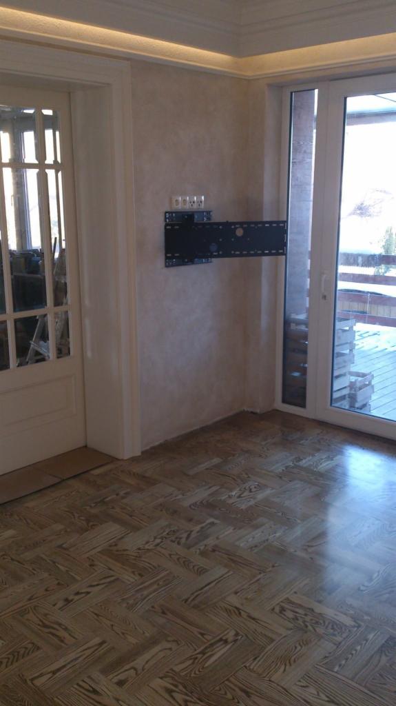 Ułożony parkiet w jodełkę francuską w jasnym odcieniu, widoczny uchwyt do telewizora na ścianie, Pilawa. Wykończenie podłóg z naturalnego drewna.