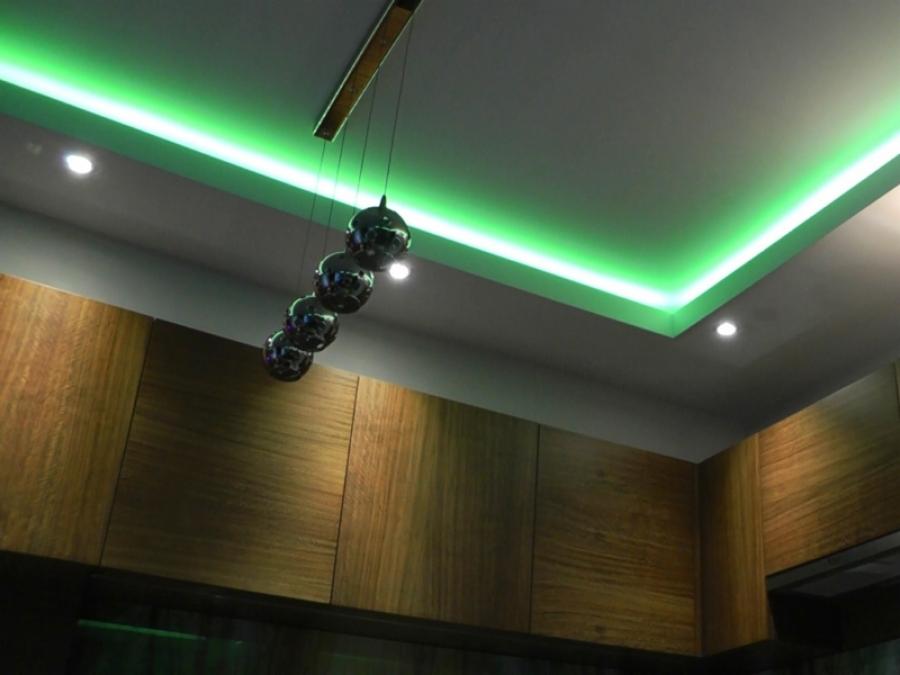 Nowoczesna kuchnia z drewnianymi szafkami, podświetlanym sufitem zielonym LED i wiszącą lampą z kulistymi kloszami. Minimalistyczny design wnętrza.