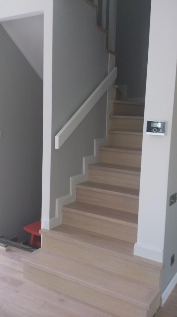 Jasne, drewniane schody z białą balustradą w szarym wnętrzu. Widoczny fragment podłogi i czerwone krzesełko w tle. Minimalistyczny design, nowoczesne wykończenie.