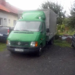 Zielony bus Volkswagen LT z plandeką na posesji, widoczny numer rejestracyjny. Tło: dom z czerwoną dachówką i zieleń.