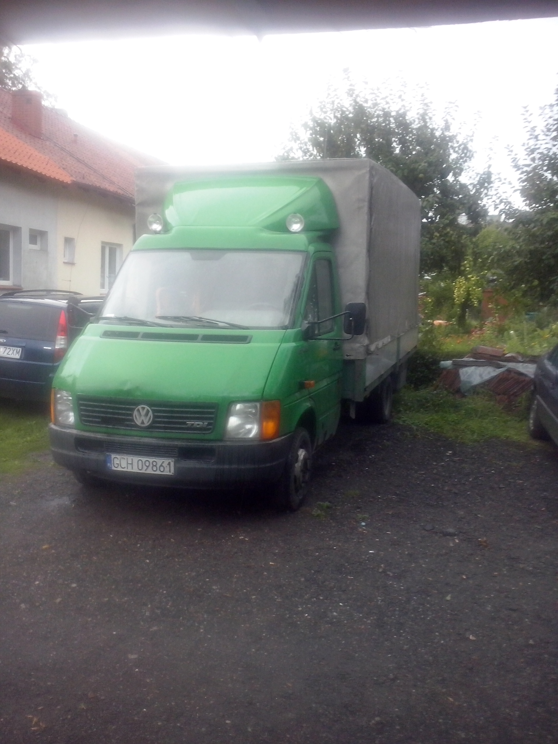 Zielony bus Volkswagen LT z plandeką na posesji, widoczny numer rejestracyjny. Tło: dom z czerwoną dachówką i zieleń.