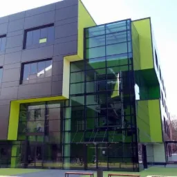 Nowoczesny, trzypiętrowy budynek z szarymi panelami i elementami w odcieniach zieleni, z dużą, przeszkloną fasadą. Na pierwszym planie ławka i rowery.