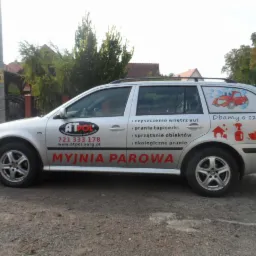 Srebrny samochód kombi z reklamą myjni parowej ATPOL, z hasłem 'Dbamy o czystość' i ikonami usług czyszczenia wnętrz, tapicerki i obiektów. Widok z boku na tle zieleni.