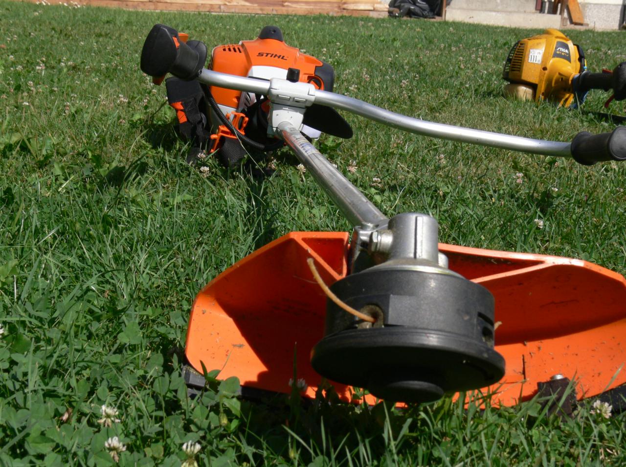 Pomarańczowa kosa spalinowa Stihl na trawie, z widoczną głowicą tnącą i fragmentem innej kosy w tle. Sprzęt ogrodniczy na trawniku.