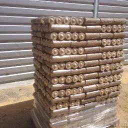 Ułożony na palecie, owinięty folią stos brykietu drzewnego na tle blaszanej ściany w słoneczny dzień. Widoczne końcówki brykietu.