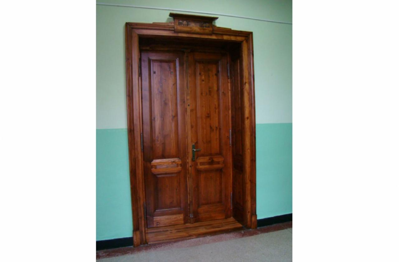 Odnowione, dwuskrzydłowe drzwi drewniane w ciemnym odcieniu brązu, z dekoracyjnymi elementami i futryną, na tle ściany pomalowanej dwukolorowo (zielony i biały).