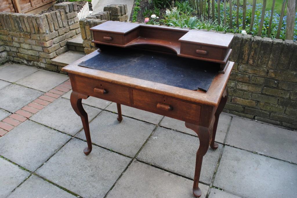 Antyczny drewniany sekretarzyk z dwoma szufladami na blacie i dwoma pod blatem, stojący na zewnątrz na kamiennej posadzce, z fragmentem ceglanego murku w tle.