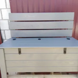 Drewniana skrzynia z siedziskiem i oparciem, pomalowana na szaro, z metalowymi zawiasami, na tle czerwonej blachy i białych pustaków.