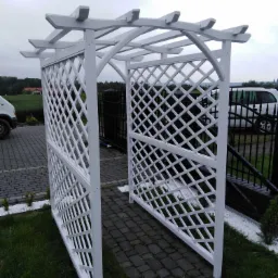 Biała, drewniana konstrukcja z kratownicą, tworząca przejście w ogrodzie, obok kostki brukowej i trawnika z białymi kamykami dekoracyjnymi.