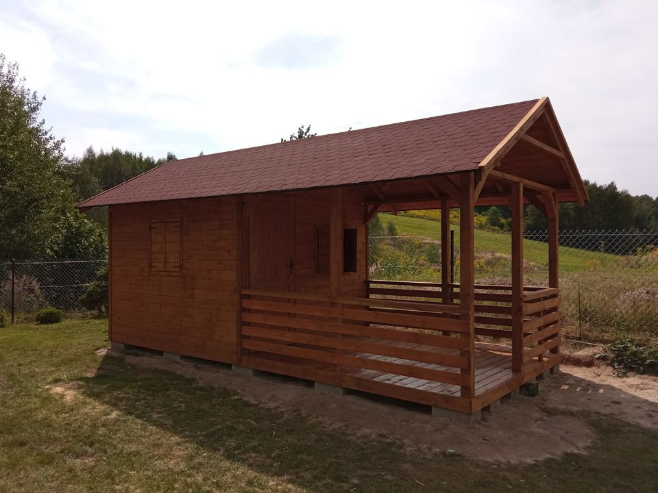 Brązowa drewniana konstrukcja z czerwonym dachem z gontów bitumicznych, częściowo zamknięta, z otwartą przestrzenią tarasową, w ogrodzie z widokiem na las w tle.