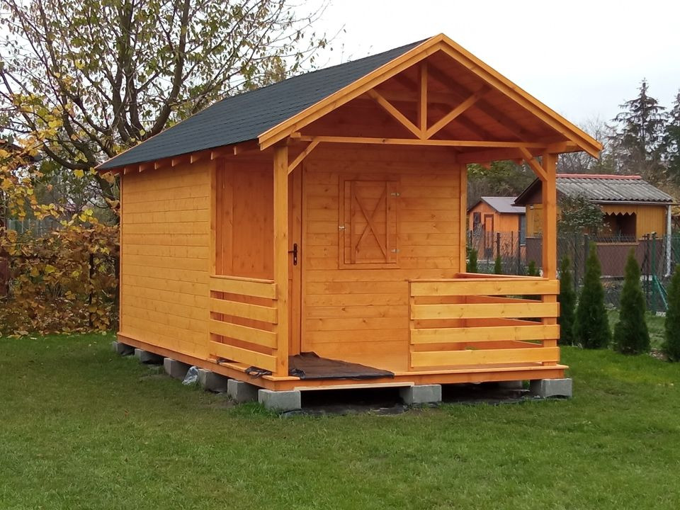 Mała, drewniana konstrukcja z czarnym dachem, podniesiona na betonowych blokach, otoczona zielonym trawnikiem i drzewami.