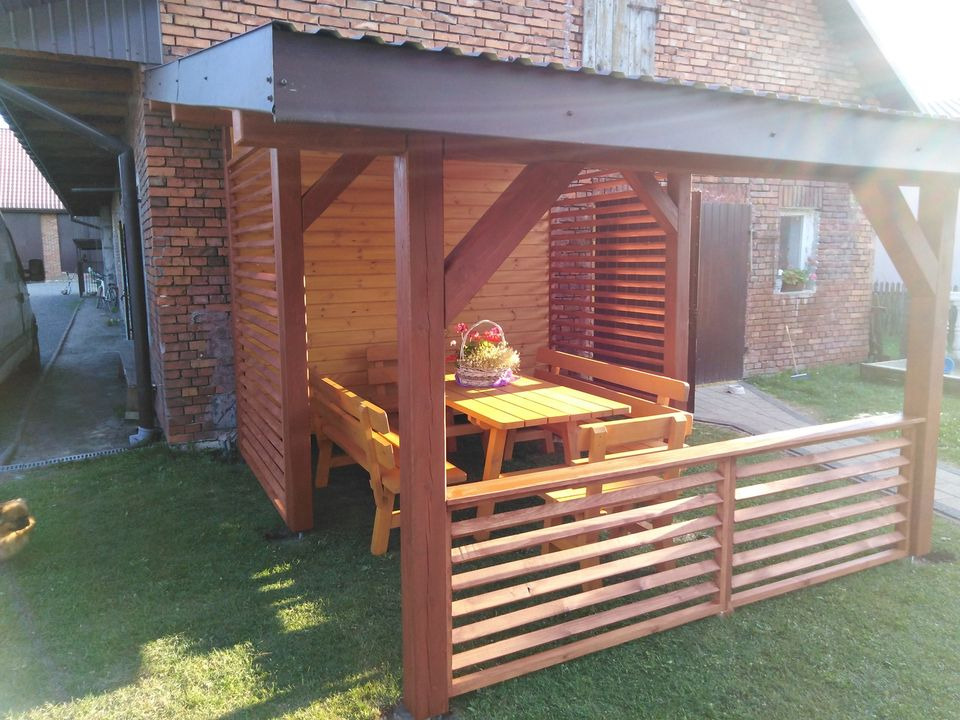 Drewniana konstrukcja zadaszona blachą, z ażurowymi ściankami i balustradą, wyposażona w stół i ławki, na trawniku przy ceglanym budynku.