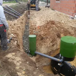 Montaż przydomowej oczyszczalni ścieków: wykop z dwoma zielonymi zbiornikami, szarymi rurami i pracownikiem z łopatą w tle, widoczny mini-koparka i fragment budynku w trakcie budowy.