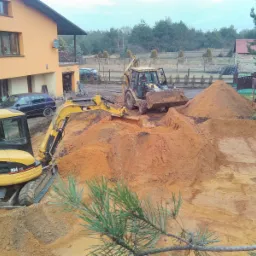 Żółty minikoparka CAT 304 CR i ładowarka pracują na placu budowy, przygotowując teren pod fundamenty; w tle dom jednorodzinny z balkonem i samochody.