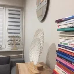 Fragment pokoju z drewnianym zegarem na ścianie, dekoracyjną, ażurową rzeźbą w kształcie liścia na drewnianej komodzie oraz stosem książek z widocznymi tytułami, neutralna kolorystyka.