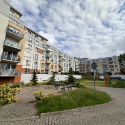 Nowoczesne budynki wielorodzinne z balkonami, widok na zagospodarowany teren zielony z ławkami i placem zabaw dla dzieci, jasne niebo z chmurami w tle.