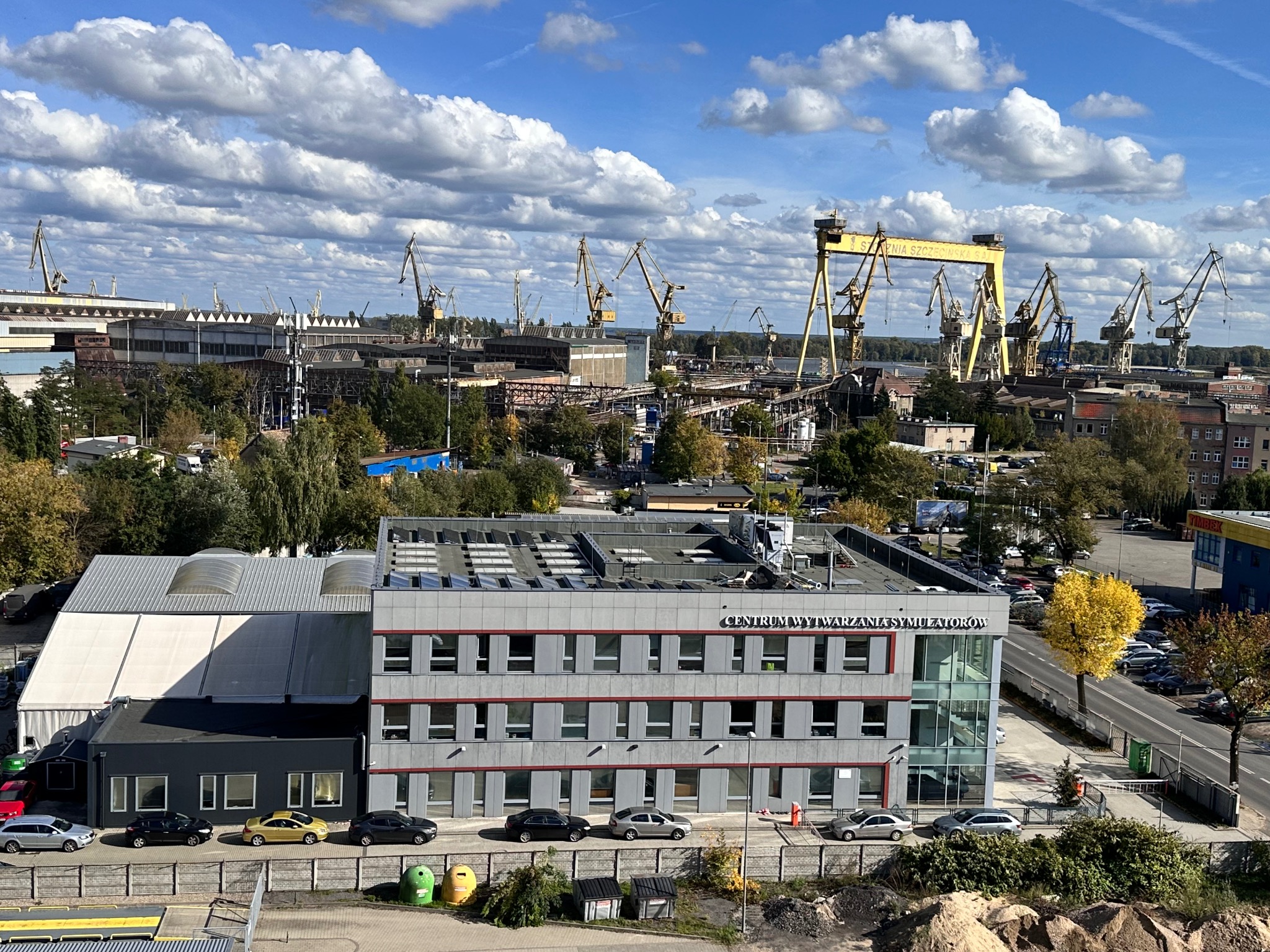 Widok na teren stoczni z licznymi dźwigami, budynkami przemysłowymi i biurowcem z napisem 'Centrum Wytwarzania Symulatorów' w słoneczny dzień z chmurami.