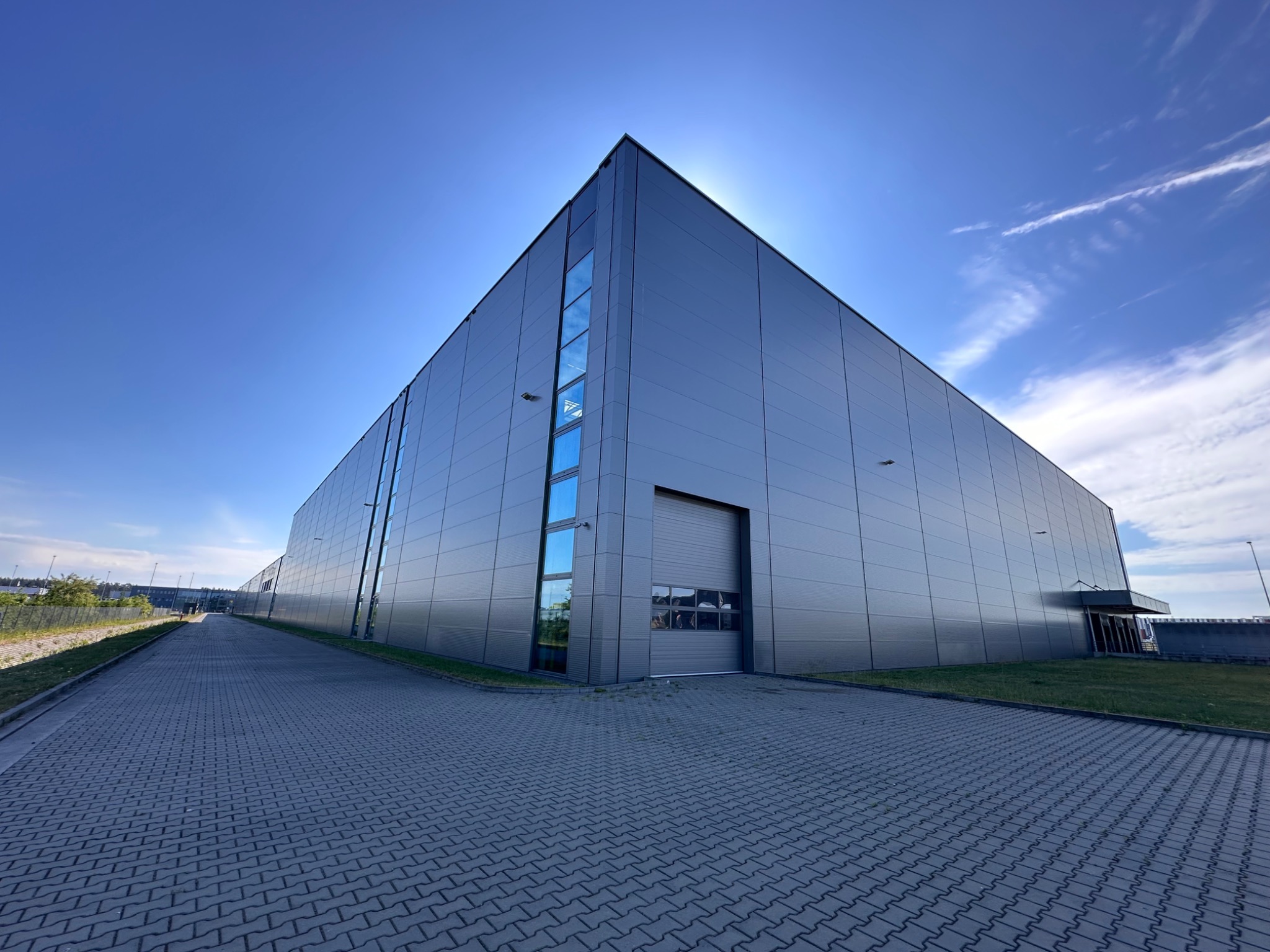 Nowoczesny, szary budynek magazynowy z panelami, z oknami pionowymi i bramą garażową, sfotografowany w słoneczny dzień z perspektywy żabiej, z widoczną kostką brukową i fragmentem nieba z chmurami.