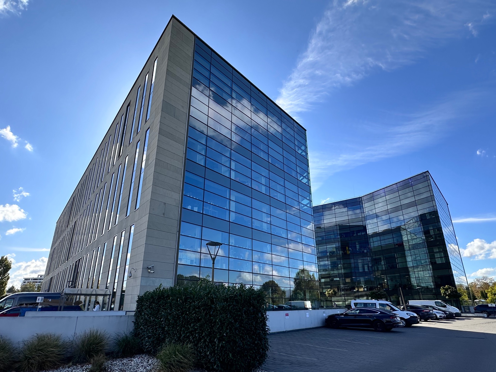 Nowoczesny budynek biurowy z fasadą ze szkła i betonu, z widocznymi odbiciami chmur na szklanej powierzchni, zaparkowane samochody przed budynkiem.