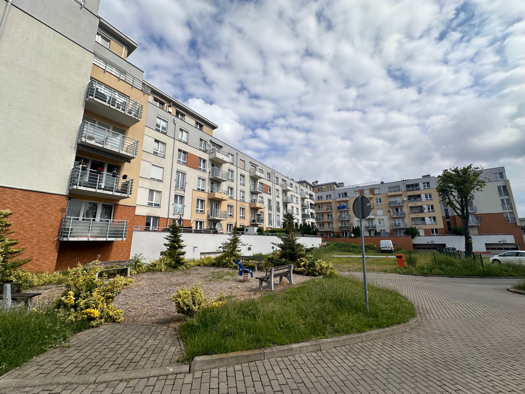 Nowoczesne budynki wielorodzinne z balkonami, widok na zagospodarowany teren zielony z ławkami i placem zabaw dla dzieci, jasne niebo z chmurami w tle.