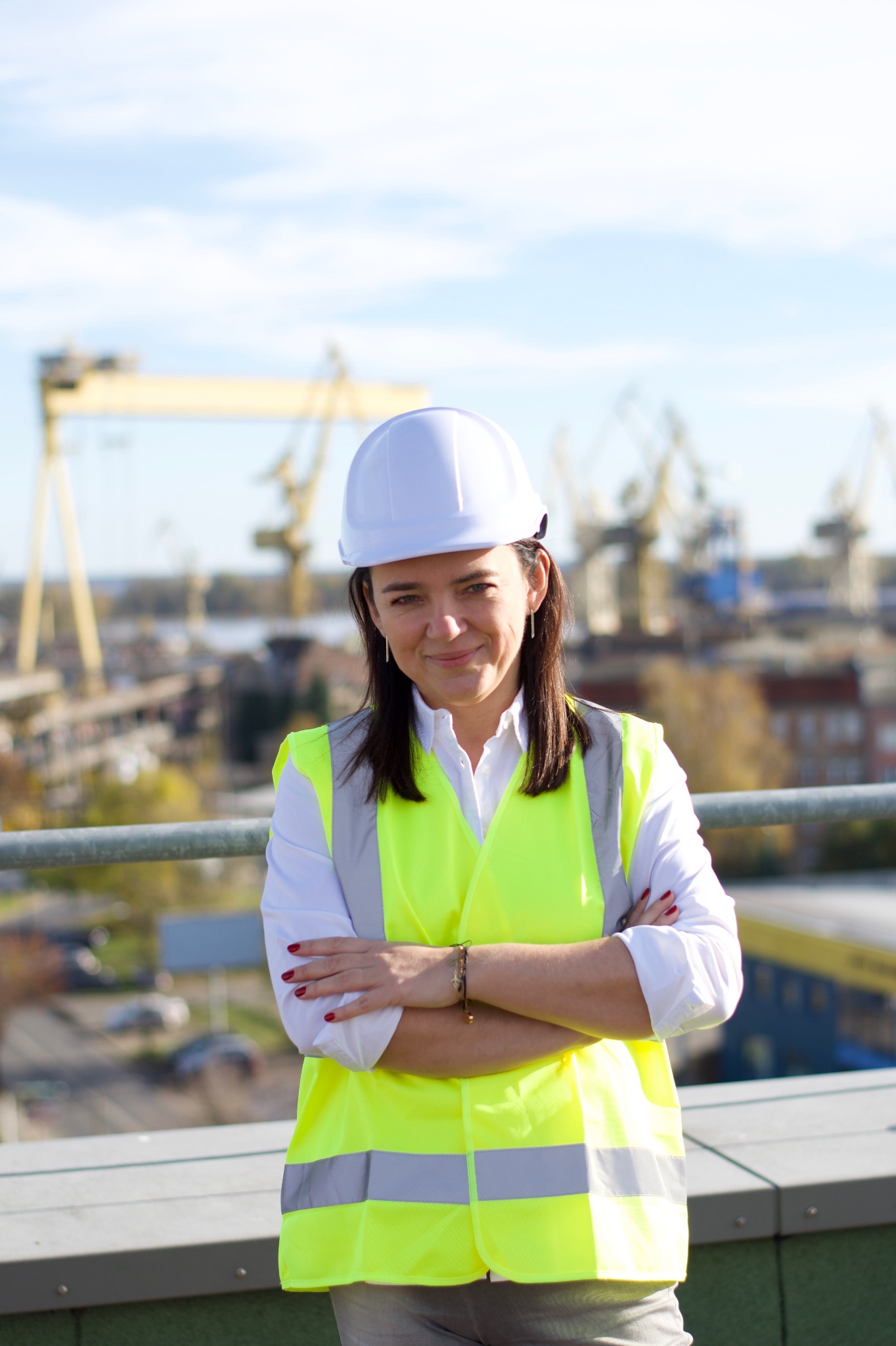 Kobieta w białym kasku i żółtej kamizelce odblaskowej, z założonymi rękami, na tle budowy z dźwigami portowymi w oddali.