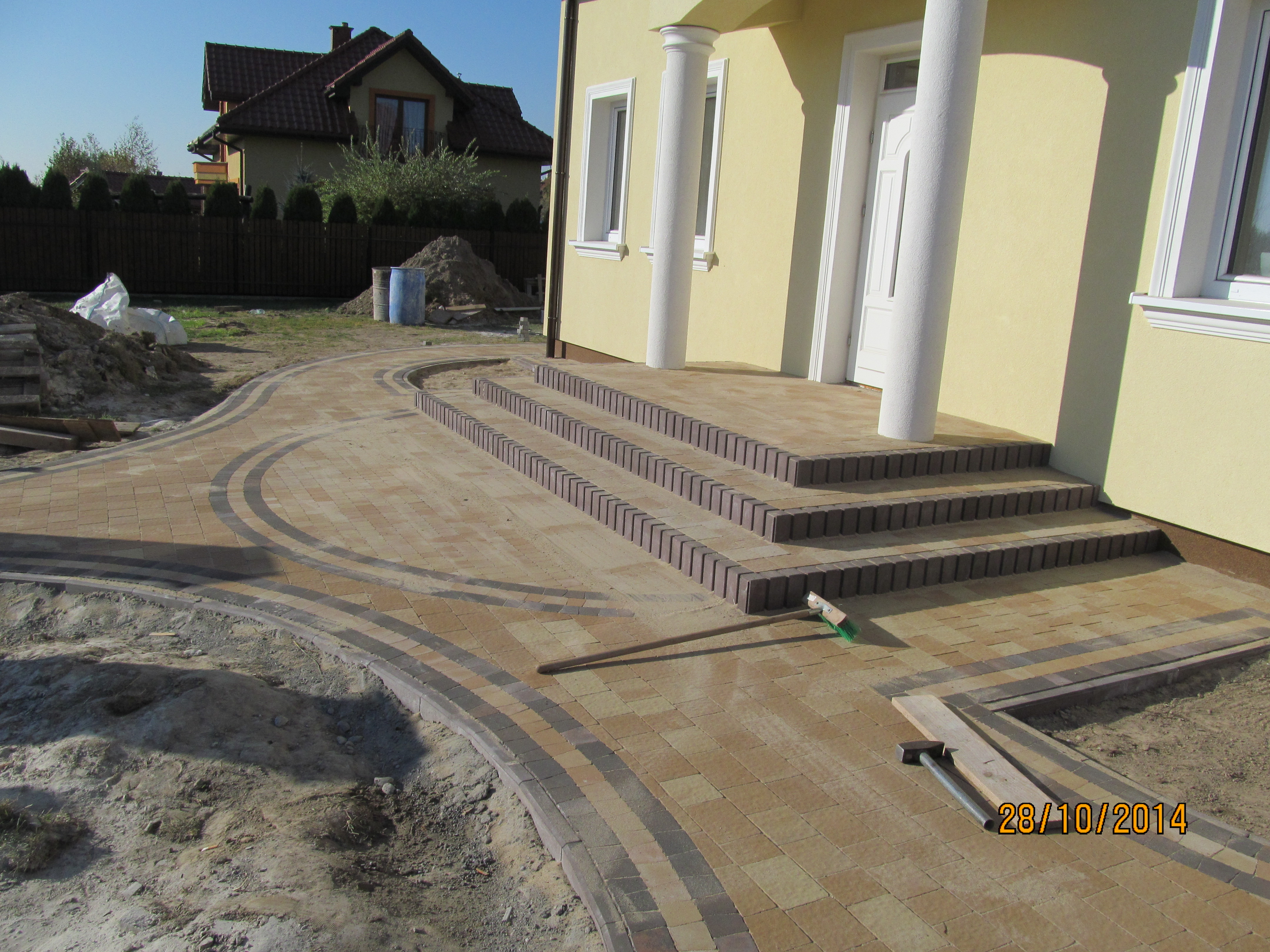 Ułożona ścieżka z kostki brukowej w odcieniach beżu i szarości prowadząca do schodów wejściowych domu z kolumnami, widoczna szczotka i fragment narzędzi brukarskich.