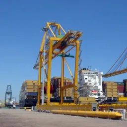 Żółty suwnicowy dźwig portowy z kontenerami na tle statku i błękitnego nieba. Dźwig na nabrzeżu z widocznymi elementami konstrukcji stalowej.