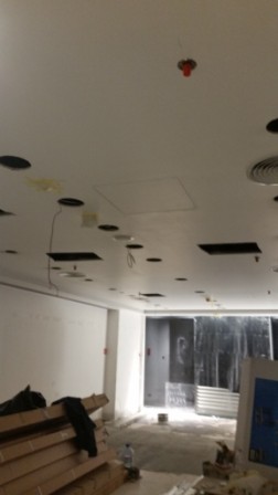 Wnętrze w trakcie prac wykończeniowych: sufit z otworami na oświetlenie, widoczne instalacje, ściany przygotowane do malowania. W tle materiały budowlane.