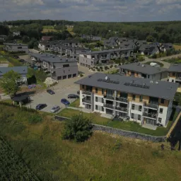 Nowoczesne osiedle budynków wielorodzinnych z szarymi dachami i panelami słonecznymi, widok z lotu ptaka na zagospodarowany teren z parkingiem i zielenią.