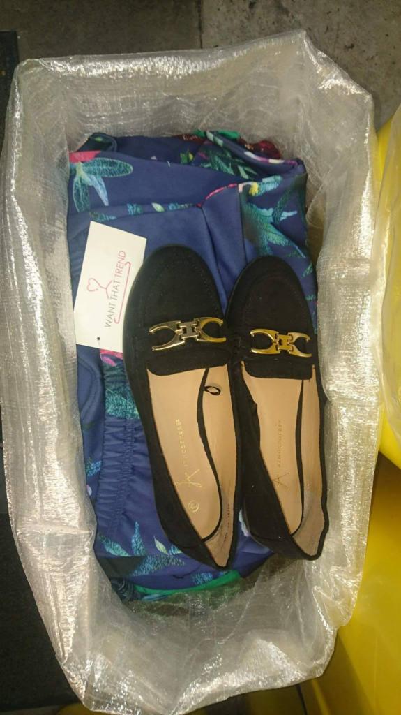 Para czarnych, zamszowych mokasynów ze złotymi zdobieniami leży na wierzchu niebieskich spodni w tropikalne wzory, całość zapakowana w przezroczystą torbę foliową z etykietą 'WANT THAT TREND'.