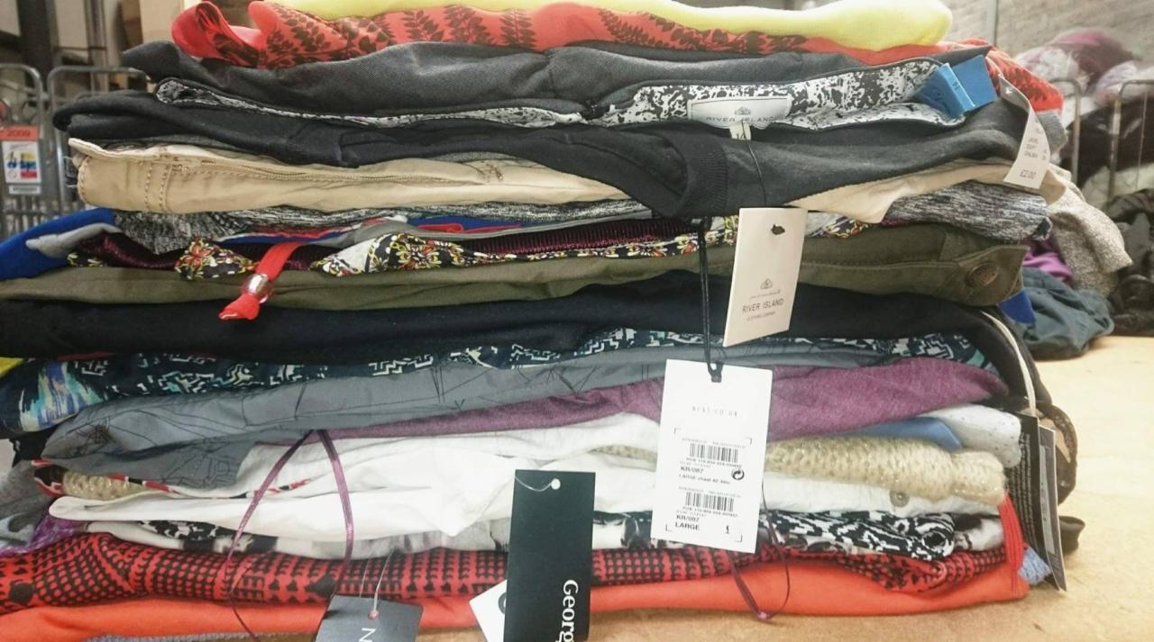 Stos ubrań z widocznymi metkami różnych marek, w tym River Island i George, ułożony na podłodze w magazynie.