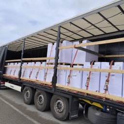 TSL ECO Sp. z o.o. - Ciężarówka z otwartą naczepą, zabezpieczona pasami transportowymi, wypełniona paletami z białymi kartonami. Widoczne koła, opony i fragment tablicy rejestracyjnej.