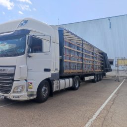 TSL ECO Sp. z o.o. - Biała ciężarówka DAF XF z naczepą Krone, załadowana paletami z szarymi, okrągłymi elementami, stoi na parkingu przed magazynem w słoneczny dzień. Widoczna drabina do naczepy.