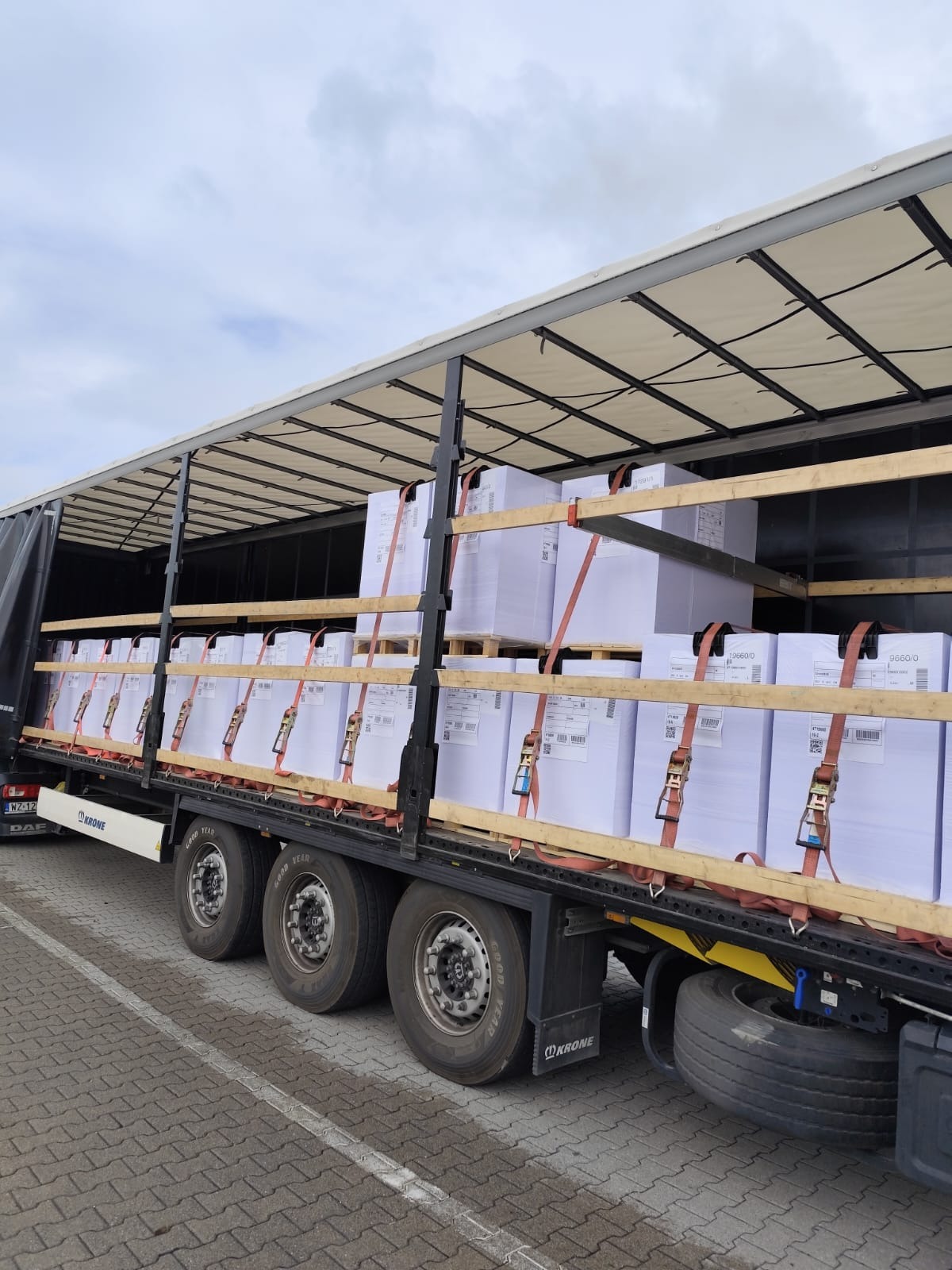 Ciężarówka z otwartą naczepą, zabezpieczona pasami transportowymi, wypełniona paletami z białymi kartonami. Widoczne koła, opony i fragment tablicy rejestracyjnej.