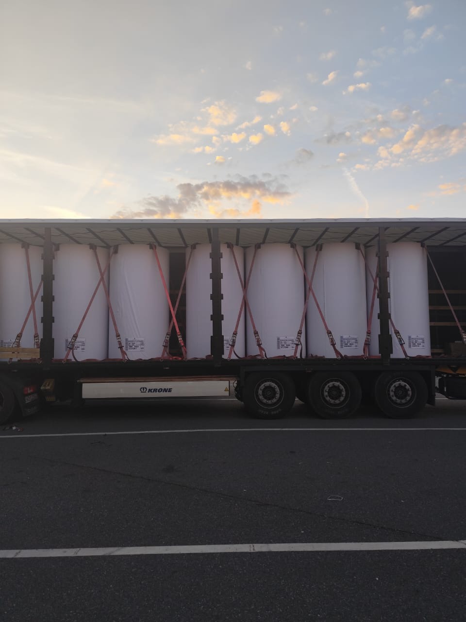 Ciężarówka marki Krone z ładunkiem zabezpieczonym czerwonymi pasami transportowymi na tle nieba o zmierzchu. Widok z boku, asfaltowa droga.