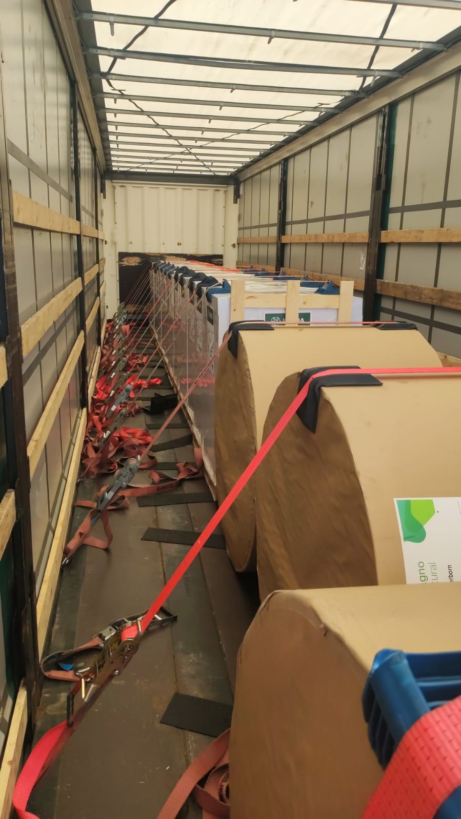 Wnętrze naczepy z ładunkiem zabezpieczonym pasami transportowymi. Widoczne rolki w kartonach i skrzynie, przygotowane do transportu. Perspektywa z głębi naczepy.