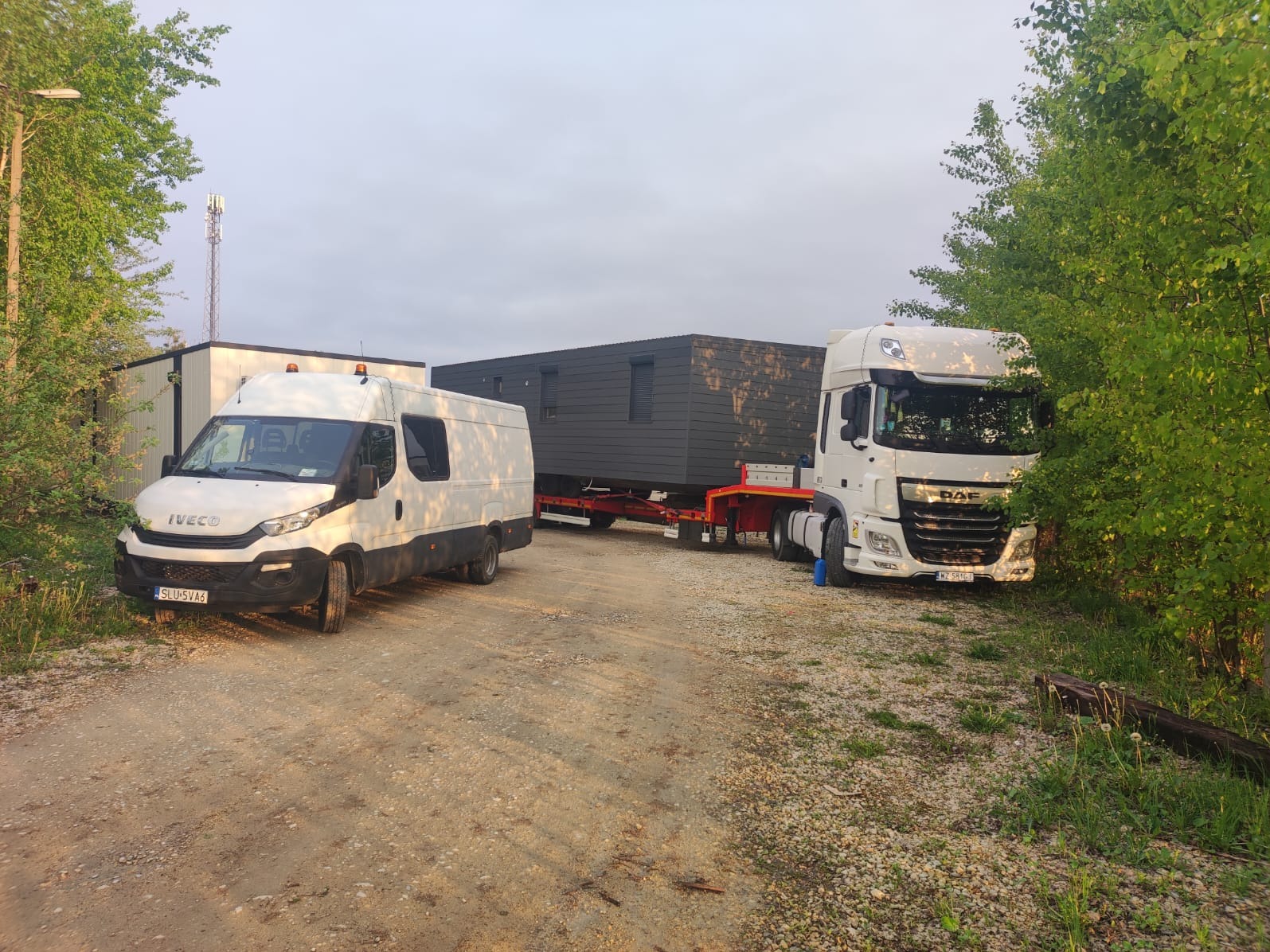 Transport modułowego domu na niskopodwoziowej lawecie, eskortowany przez busa Iveco i ciągnik DAF, na żwirowym placu w otoczeniu zieleni i budynków.