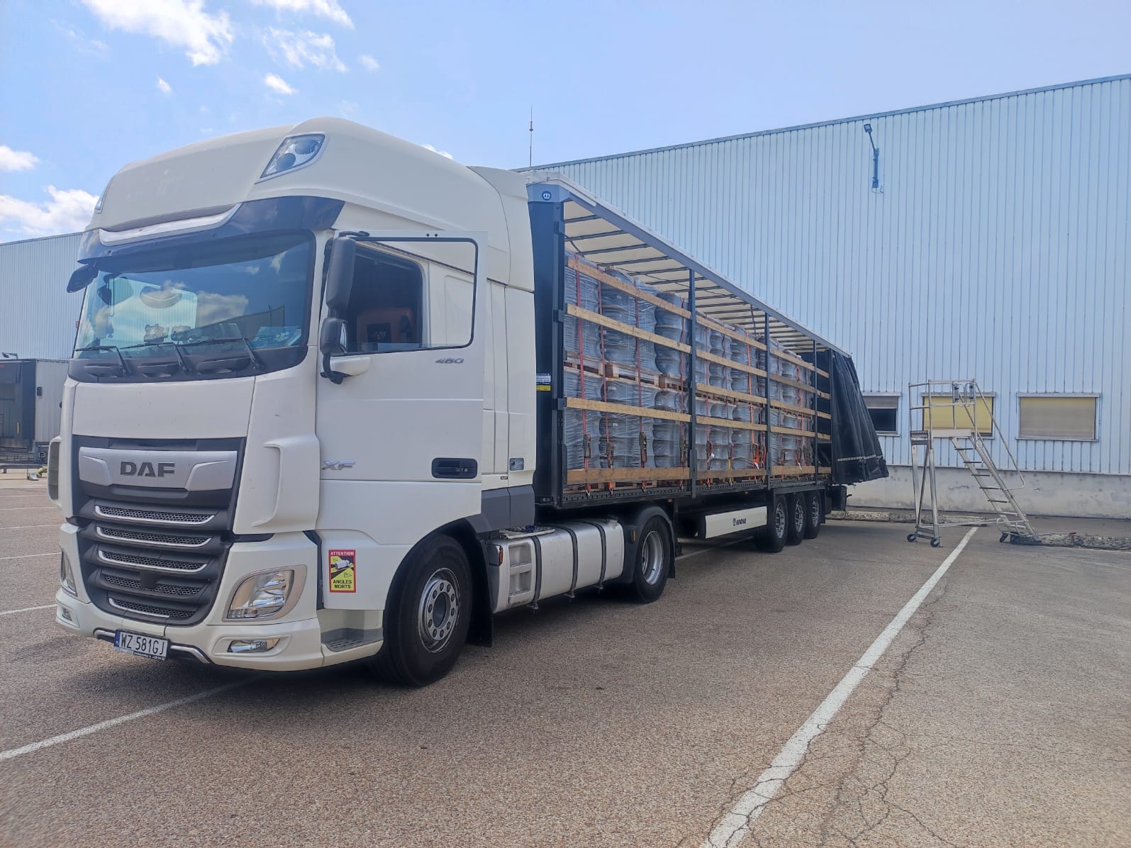 Biała ciężarówka DAF XF z naczepą Krone, załadowana paletami z szarymi, okrągłymi elementami, stoi na parkingu przed magazynem w słoneczny dzień. Widoczna drabina do naczepy.