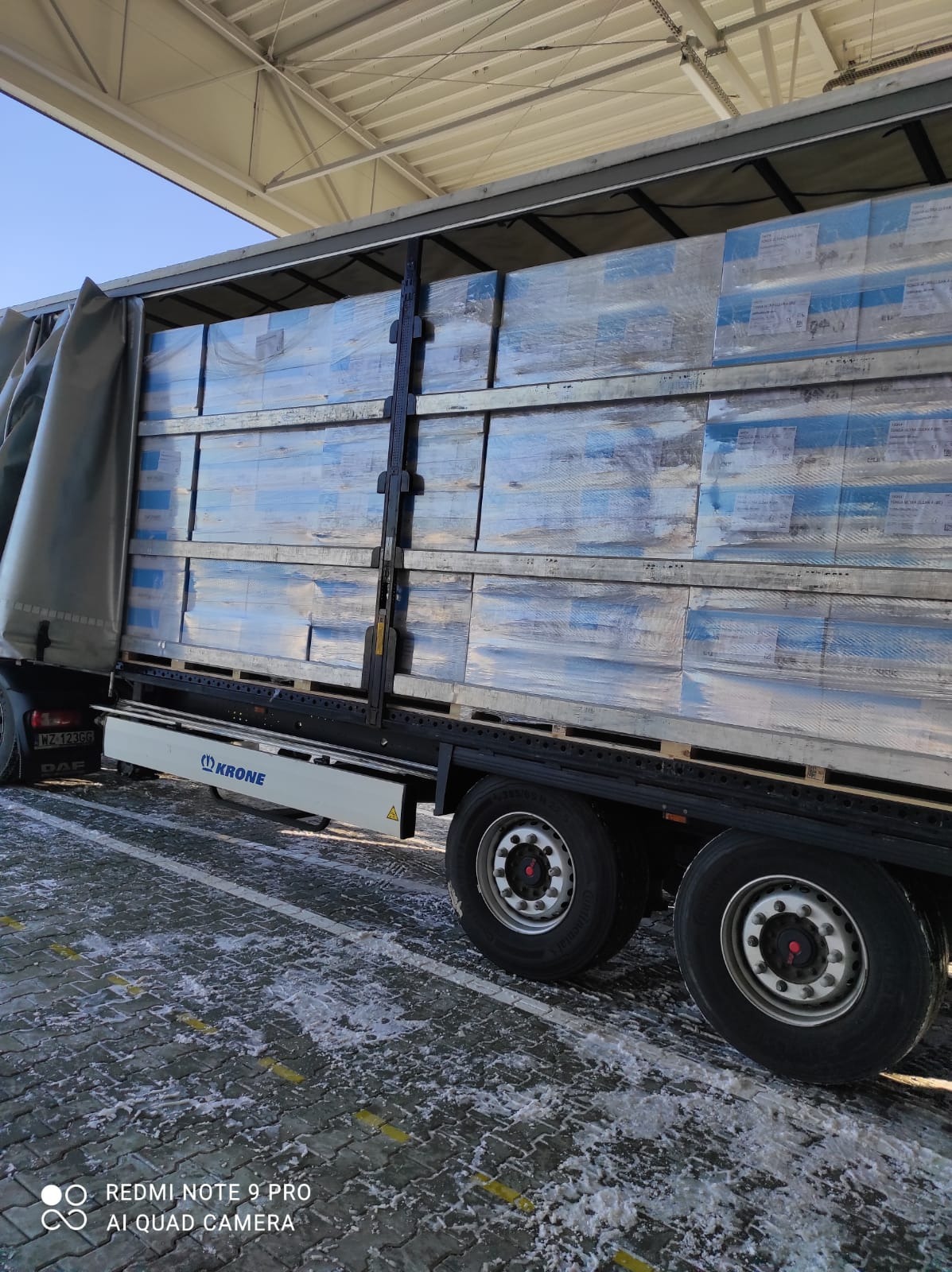 Ciężarówka DAF z naczepą Krone, załadowana paletami z towarem, zabezpieczonym folią stretch. Widoczne opony i fragment plandeki. Zimowa sceneria z resztkami śniegu na kostce brukowej.