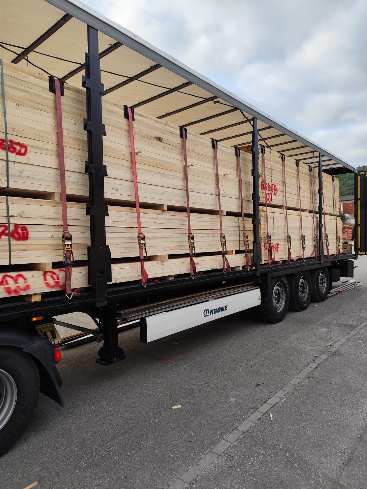 Ciężarówka marki Krone z ładunkiem drewna zabezpieczonym pasami, widoczna w Lublińcu. Transport drewna na paletach, częściowo otwarty naczepą.