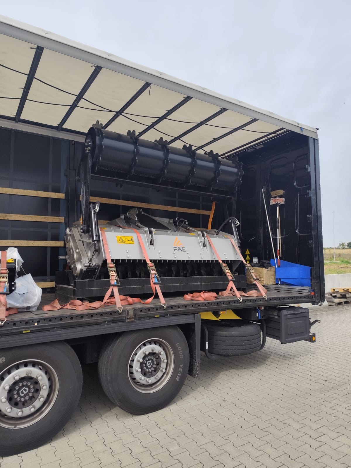 Transport specjalistycznej maszyny rolniczej FAE w naczepie ciężarówki, zabezpieczonej pasami transportowymi. Widoczne koła ciężarówki i fragment rampy załadunkowej.