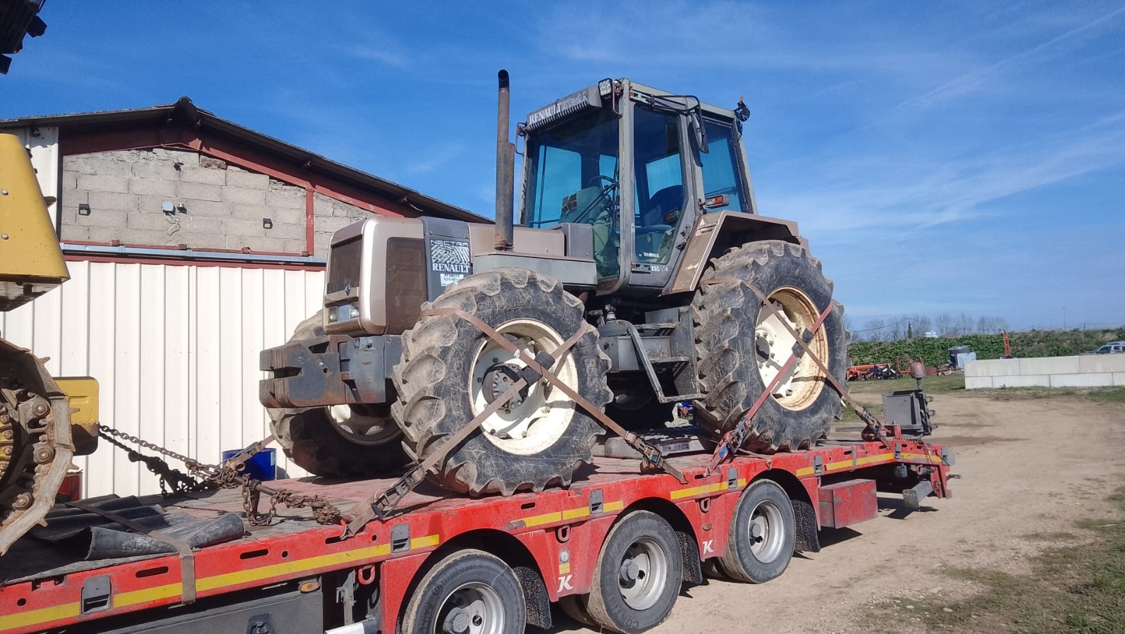 Ciężki traktor Renault na czerwonej platformie niskopodwoziowej, zabezpieczony łańcuchami. Błękitne niebo w tle, częściowo widoczny budynek gospodarczy.