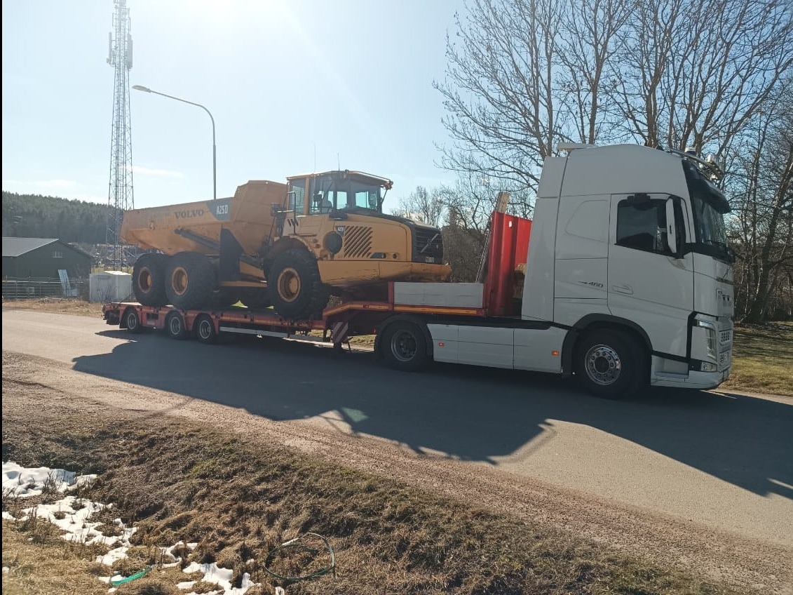 Biała ciężarówka z naczepą niskopodwoziową przewozi żółty wozidło Volvo A25D na tle wieży telekomunikacyjnej i drzew bez liści w słoneczny dzień.
