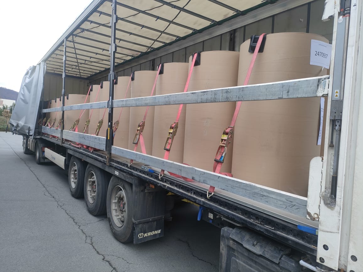 Ciężarówka z otwartą plandeką, załadowana rolami papieru zabezpieczonymi pasami transportowymi. Widoczne tabliczki z numerami identyfikacyjnymi na rolach.
