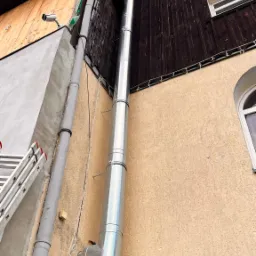 Zewnętrzny, metalowy przewód spalinowy zamocowany do ściany budynku, z widoczną drabiną opartą o ścianę i kamerą monitoringu pod dachem.