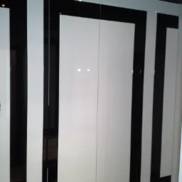 Biała szafa wnękowa z czarnymi, geometrycznymi obramowaniami na frontach drzwi przesuwnych, oświetlenie punktowe na suficie.