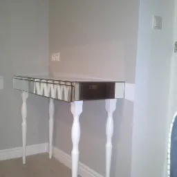 Elegancka toaletka z lustrzanym blatem i białymi, toczonymi nogami, ustawiona przy szarej ścianie z gniazdkiem i białymi drzwiami. Minimalistyczny design.