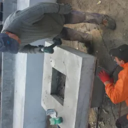 Dwóch robotników pracuje nad betonowymi elementami konstrukcyjnymi z otworem, używając wiertarki i szpachli, na placu budowy z widocznymi kamieniami i zielenią w tle.